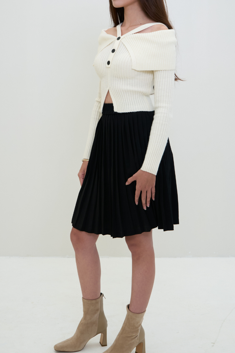 Women wearing a white cropped cardigan over a black pleated skirt with beige ankle boots on a white background