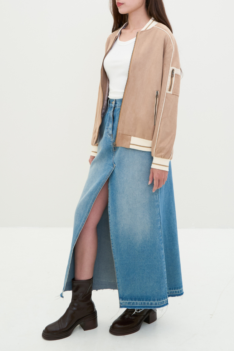 Woman wearing a beige jacket, white top, and blue denim skirt on a light gray background. Person wearing a beige bomber jacket with white trim. NAILA BASIC jacket in beige color with zipper. Unisex jacket. Full body styling image.