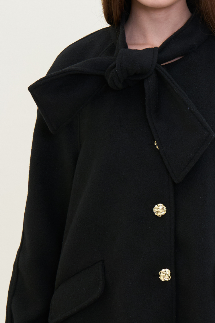 Black coat with a large bow and gold buttons on a neutral background. Black coat with fur cuffs and gold buttons. NOOR coat Women’s back coat. Wool, Combined Cotton, Vegan Fur. Close up image.